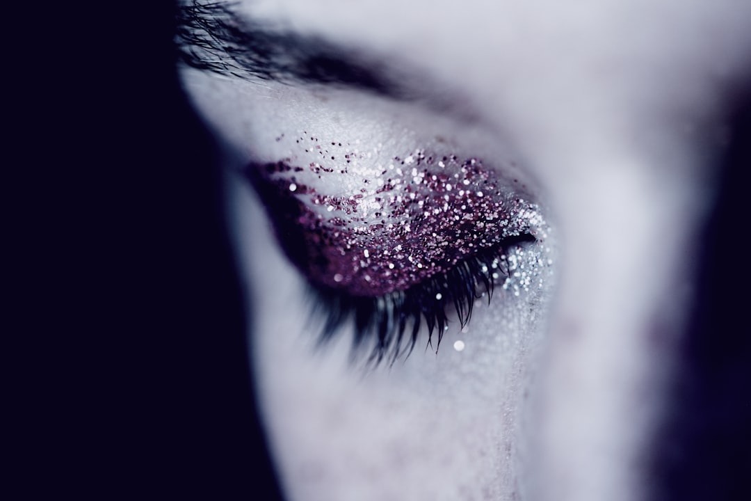 A close-up of a closed eye with long eyelashes and eyelid covered in shimmering pink and silver glitter makeup, set against a soft, out-of-focus background.