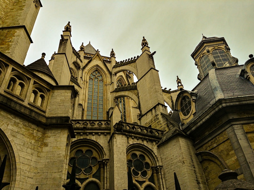 A dramatic upward view of a Gothic cathedral with pointed arches, tall stained glass windows, and decorative stonework under a cloudy sky.