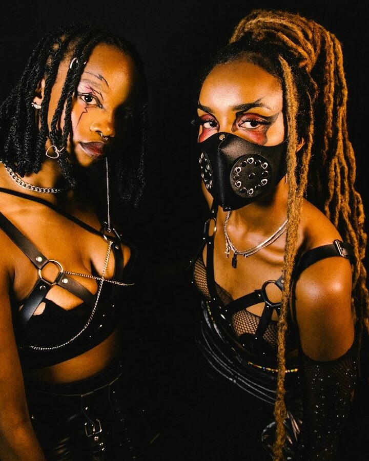 Two people in black strappy outfits and bold makeup pose against a dark background. One has long light brown dreadlocks and wears a black mask with metal rings; the other has short twists and a nose ring.