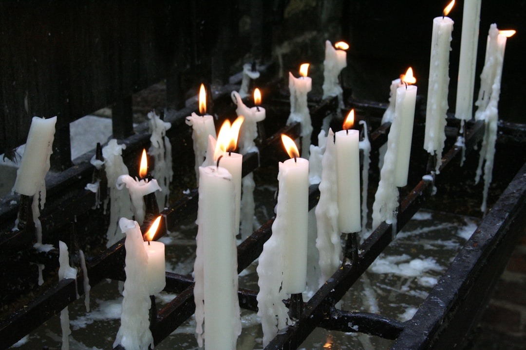 Several white candles, some partially melted, stand upright on a black metal rack. Their flames flicker, and wax drips down onto the surface below, creating pools and trails of hardened wax.