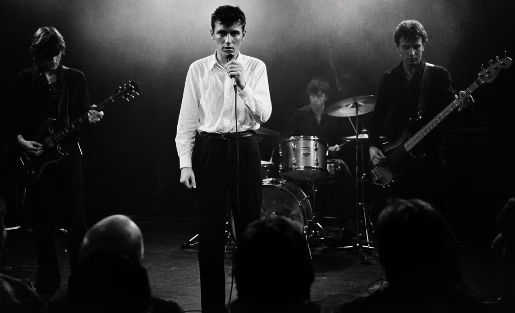 Joy Division performing live in black and white, with Ian Curtis singing at center stage as guitarist Bernard Sumner, bassist Peter Hook, and drummer Stephen Morris perform behind him.