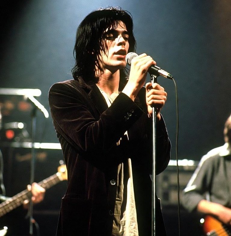 Lou Reed performing with The Velvet Underground, singing into a microphone onstage in a dark jacket with band members and instruments behind him.